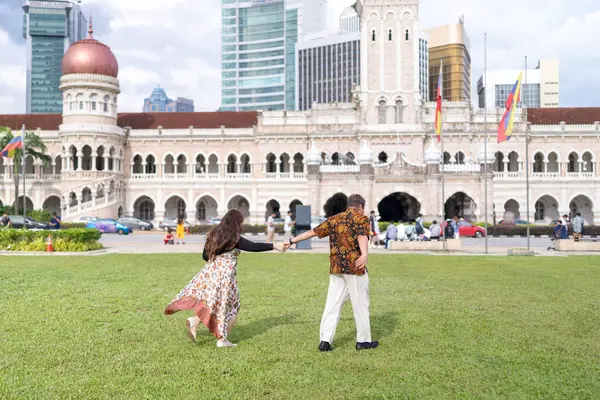 50 'li yaşlarda Malezyalı bir adam ve 30' lu yaşlarda Çinli bir kadın. İkisi de geleneksel Malezya kıyafeti giymiş. Malezya 'nın başkenti Kuala Lumpur' daki tarihi binanın önünde çimlerin üzerinde birlikte yürüyorlar..