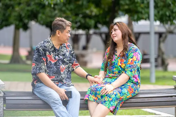 50 'li yaşlarda Malezyalı bir adam ve 30' lu yaşlarında Çinli bir kadın, ikisi de renkli giyinmiş, yakın bir bankta oturuyor ve Malezya 'nın Kuala Lumpur kentindeki KLCC Parkı' nda birlikte yürüyorlar..