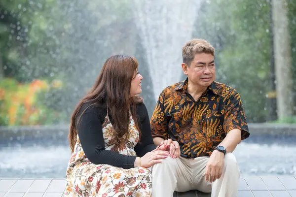 50 'li yaşlarda Malezyalı bir adam geleneksel 30' lu yaşlarda geleneksel bir elbise giyen Çinli bir kadın Kuala Lumpur 'daki Botani Perdana çeşmesinin önünde yan yana oturuyor.