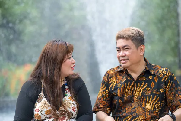 50 'li yaşlarda Malezyalı bir adam geleneksel 30' lu yaşlarda geleneksel bir elbise giyen Çinli bir kadın Kuala Lumpur 'daki Botani Perdana çeşmesinin önünde yan yana oturuyor.