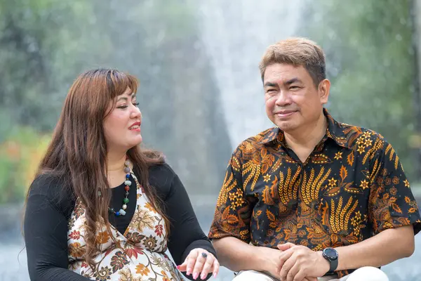 50 'li yaşlarda Malezyalı bir adam geleneksel 30' lu yaşlarda geleneksel bir elbise giyen Çinli bir kadın Kuala Lumpur 'daki Botani Perdana çeşmesinin önünde yan yana oturuyor.