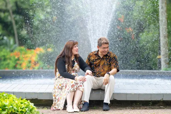 50 'li yaşlarda Malezyalı bir adam geleneksel 30' lu yaşlarda geleneksel bir elbise giyen Çinli bir kadın Kuala Lumpur 'daki Botani Perdana çeşmesinin önünde yan yana oturuyor.