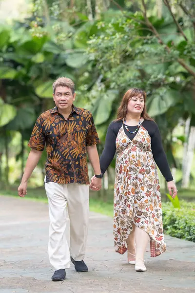 50 'li yaşlarda Malezyalı bir adam geleneksel Malezya gömleği giyiyor ve 30' lu yaşlarda Çinli bir kadın geleneksel elbiseler giyiyor, Taman Botani Perdana, Kuala Lumpur, Malezya 'da bitkilerle çevrili yürüyor..