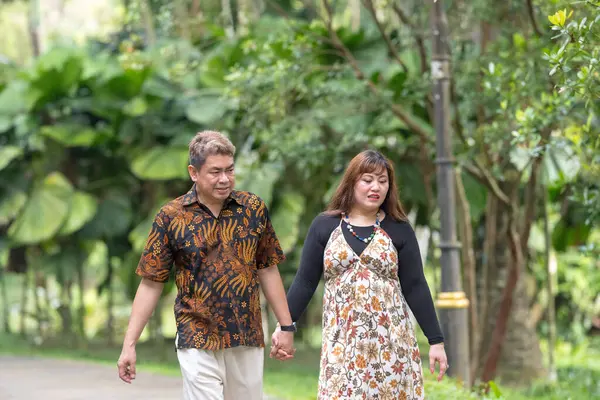 50 'li yaşlarda Malezyalı bir adam geleneksel Malezya gömleği giyiyor ve 30' lu yaşlarda Çinli bir kadın geleneksel elbiseler giyiyor, Taman Botani Perdana, Kuala Lumpur, Malezya 'da bitkilerle çevrili yürüyor..