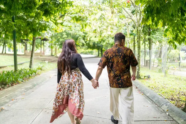 50 'li yaşlarda Malezyalı bir adam geleneksel Malezya gömleği giyiyor ve 30' lu yaşlarda Çinli bir kadın geleneksel elbiseler giyiyor, Taman Botani Perdana, Kuala Lumpur, Malezya 'da bitkilerle çevrili yürüyor..