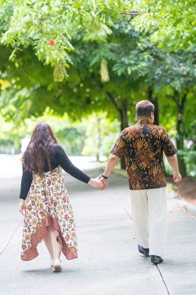 50 'li yaşlarda Malezyalı bir adam geleneksel Malezya gömleği giyiyor ve 30' lu yaşlarda Çinli bir kadın geleneksel elbiseler giyiyor, Taman Botani Perdana, Kuala Lumpur, Malezya 'da bitkilerle çevrili yürüyor..