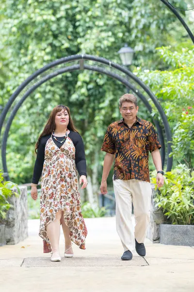 50 'li yaşlarda Malezyalı bir adam geleneksel Malezya gömleği giyiyor ve 30' lu yaşlarda Çinli bir kadın geleneksel elbiseler giyiyor, Taman Botani Perdana, Kuala Lumpur, Malezya 'da bitkilerle çevrili yürüyor..