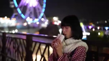 Taipei, Shilin Bölgesi 'nde yirmili yaşlarında Tayvanlı bir kadın Tayvan' daki lunaparkın arka planında güzel aydınlatılmış dönme dolap ile taze patates kızartması yiyor..