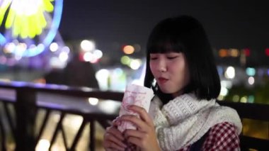 Taipei, Shilin Bölgesi 'nde yirmili yaşlarında Tayvanlı bir kadın Tayvan' daki lunaparkın arka planında güzel aydınlatılmış dönme dolap ile taze patates kızartması yiyor..