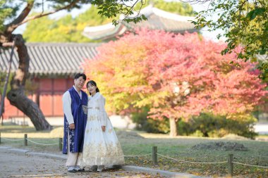 30 'lu yaşlarda Koreli bir çift ve 20' li yaşlarda bir kadın mutlu bir şekilde Hanbok giyiyor ve Güney Kore 'nin Seul kentindeki tarihi bir binada güzel sonbahar yapraklarının önünde vakit geçiriyorlar..