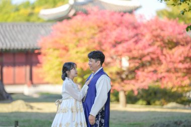 30 'lu yaşlarda Koreli bir çift ve 20' li yaşlarda bir kadın mutlu bir şekilde Hanbok giyiyor ve Güney Kore 'nin Seul kentindeki tarihi bir binada güzel sonbahar yapraklarının önünde vakit geçiriyorlar..