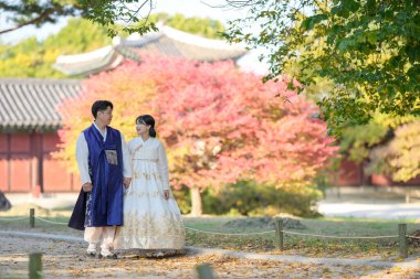 30 'lu yaşlarda Koreli bir çift ve 20' li yaşlarda bir kadın mutlu bir şekilde Hanbok giyiyor ve Güney Kore 'nin Seul kentindeki tarihi bir binada güzel sonbahar yapraklarının önünde vakit geçiriyorlar..