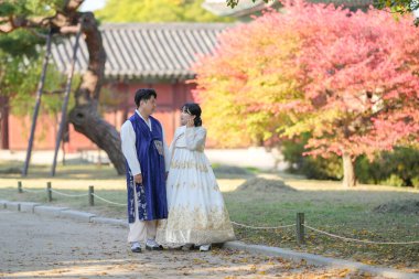 30 'lu yaşlarda Koreli bir çift ve 20' li yaşlarda bir kadın mutlu bir şekilde Hanbok giyiyor ve Güney Kore 'nin Seul kentindeki tarihi bir binada güzel sonbahar yapraklarının önünde vakit geçiriyorlar..