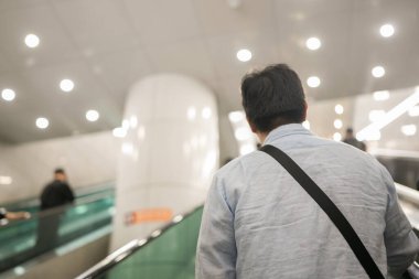 Mavi tişörtlü ve siyah sırt çantalı bir adam yürüyen merdivenden iniyor. Arka planda başkaları da var.