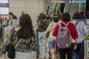 Bir grup insan havaalanı terminalinden geçiyor. Bir kadın sırt çantası takıyor. Arka planda bavul taşıyan birkaç kişi var.