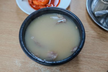 İçinde et olan bir kase çorba. Çorba beyaz ve kase siyah. Kasede bir kaşık var.