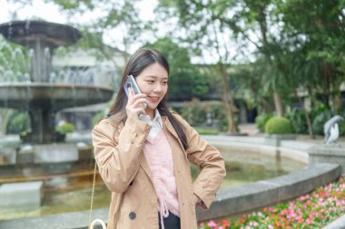 20 'li yaşlarda Tayvan' da uzun saçlı, bej renkli trençkot giyen bir kadın 30 Ocak 2024 'te öğlen vakti Tayvan' ın Taipei şehrinde Xinyi İlçesi 'ndeki bir parkta bir çeşme etrafında akıllı telefon kullanıyor..