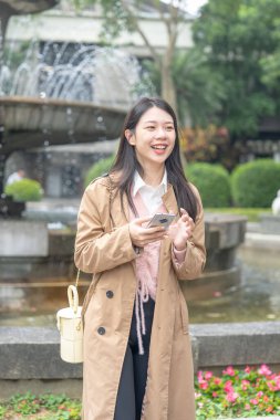 A long-haired Taiwanese woman in her 20s wearing a beige trench coat operates a smartphone around a fountain in a park in Xinyi District, Taipei City, Taiwan at noon in winter on January 30, 2024.