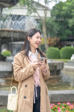 20 'li yaşlarda Tayvan' da uzun saçlı, bej renkli trençkot giyen bir kadın 30 Ocak 2024 'te öğlen vakti Tayvan' ın Taipei şehrinde Xinyi İlçesi 'ndeki bir parkta bir çeşme etrafında akıllı telefon kullanıyor..