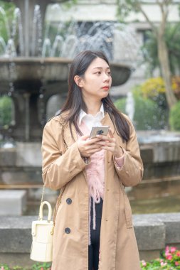20 'li yaşlarda Tayvan' da uzun saçlı, bej renkli trençkot giyen bir kadın 30 Ocak 2024 'te öğlen vakti Tayvan' ın Taipei şehrinde Xinyi İlçesi 'ndeki bir parkta bir çeşme etrafında akıllı telefon kullanıyor..