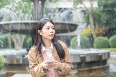 20 'li yaşlarda Tayvan' da uzun saçlı, bej renkli trençkot giyen bir kadın 30 Ocak 2024 'te öğlen vakti Tayvan' ın Taipei şehrinde Xinyi İlçesi 'ndeki bir parkta bir çeşme etrafında akıllı telefon kullanıyor..