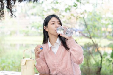 20 'li yaşlarda, Tayvan' ın Xinyi semtinde bir bankta pembe kazak giyen uzun saçlı Tayvanlı bir kadın 30 Ocak 2024 'te öğleden sonra bir plastik şişeden su içiyor..