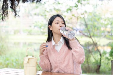 20 'li yaşlarda, Tayvan' ın Xinyi semtinde bir bankta pembe kazak giyen uzun saçlı Tayvanlı bir kadın 30 Ocak 2024 'te öğleden sonra bir plastik şişeden su içiyor..