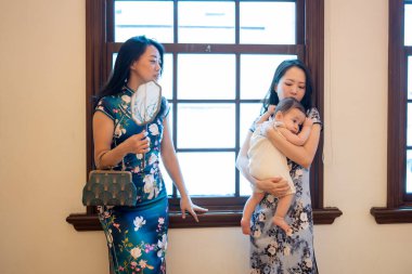 Two Taiwanese women in their 30s wearing traditional Chinese clothing hold 6-month-old baby and chat at table by the window of traditional building on Dihua Street in Taipei City, in September. clipart