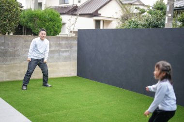 Japon bir ebeveyn ve çocuk, 20 'li yaşlarda bir adam ve 6 yaşında bir kız, evlerinin bahçesinde birbirlerine yeşil plastik toplar atarak oynuyorlar..