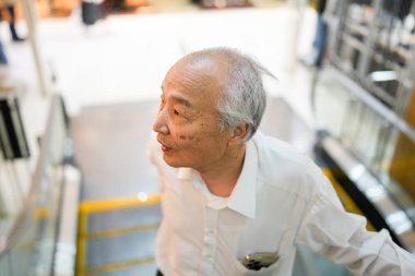 Japonya 'da kapalı bir yürüyen merdivende, 70' li yaşlarının sonlarında, temiz beyaz bir gömlek giyen bir Japon, sakin bir tavırla yürüyen merdivene biniyor,.