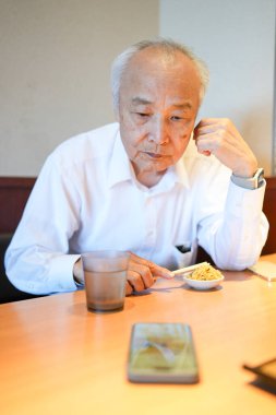 Geleneksel bir Japon lokantasında, 70 'li yaşların sonlarında beyaz gömlekli bir Japon, ahşap bir masada oturur, pirinç ve miso çorbasıyla servis edilen klasik Japon yemeklerinin tadını çıkarır..