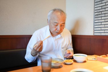 Geleneksel bir Japon lokantasında, 70 'li yaşların sonlarında beyaz gömlekli bir Japon, ahşap bir masada oturur, pirinç ve miso çorbasıyla servis edilen klasik Japon yemeklerinin tadını çıkarır..