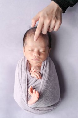 Yumuşak bir kundağa sarılmış, açık mor bir arka planda huzur içinde uyuyan iki haftalık bir Japon bebeğin profesyonel yeni doğan fotoğrafı. Bebek, anne ve babasının sıcak ellerinden nazikçe etkilenir..
