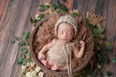 Bej örgü tulumlu, uyumlu bir şapka takan, ahşap bir güvertede kahverengi bir kürk sepetinde uyuyan iki haftalık bir bebeğin yeni doğan fotoğrafı. Profesyonel yeni doğan fotoğrafçılık..
