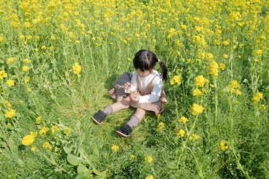 6 aylık bir bebek ve 3 yaşındaki Japon bir kız çocuğu, sakin bir kırsal bölgedeki çiçek tarlasında birlikte oturup Mart ayının sıcağında sarı çiçeklerle çevrili atıştırmalıkların tadını çıkarıyorlar..