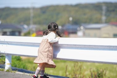 3 yaşında bir Japon kız beyaz bir korkuluğun yanında duruyor, sessizce kırlara bakıyor. Mart ayı başlarında yakalanan dişi, sakin bahar melteminin ve kırsal bir ortamda hafif güneş ışığının tadını çıkarıyor..