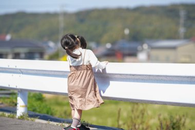 3 yaşında bir Japon kız beyaz bir korkuluğun yanında duruyor, sessizce kırlara bakıyor. Mart ayı başlarında yakalanan dişi, sakin bahar melteminin ve kırsal bir ortamda hafif güneş ışığının tadını çıkarıyor..