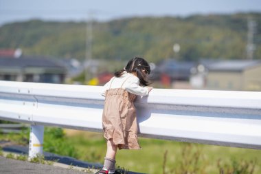 3 yaşında bir Japon kız beyaz bir korkuluğun yanında duruyor, sessizce kırlara bakıyor. Mart ayı başlarında yakalanan dişi, sakin bahar melteminin ve kırsal bir ortamda hafif güneş ışığının tadını çıkarıyor..