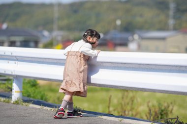 3 yaşında bir Japon kız beyaz bir korkuluğun yanında duruyor, sessizce kırlara bakıyor. Mart ayı başlarında yakalanan dişi, sakin bahar melteminin ve kırsal bir ortamda hafif güneş ışığının tadını çıkarıyor..