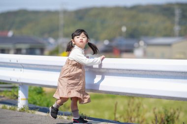 3 yaşında bir Japon kız beyaz bir korkuluğun yanında duruyor, sessizce kırlara bakıyor. Mart ayı başlarında yakalanan dişi, sakin bahar melteminin ve kırsal bir ortamda hafif güneş ışığının tadını çıkarıyor..