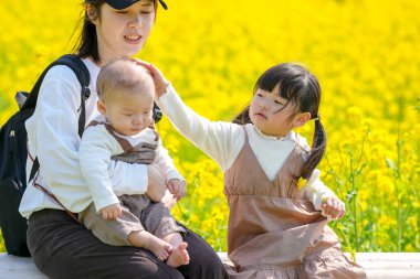 30 'lu yaşlarda Japon bir anne, kırsal bir tarlada çiçek açan bir bankta, 6 aylık bebeğini kucağında, 3 yaşındaki ikiz kuyruklu kızının yanında oturuyor. Mart ayı başlarında yakalandı..