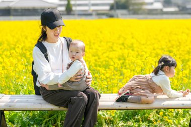 30 'lu yaşlarda Japon bir anne, kırsal bir tarlada çiçek açan bir bankta, 6 aylık bebeğini kucağında, 3 yaşındaki ikiz kuyruklu kızının yanında oturuyor. Mart ayı başlarında yakalandı..