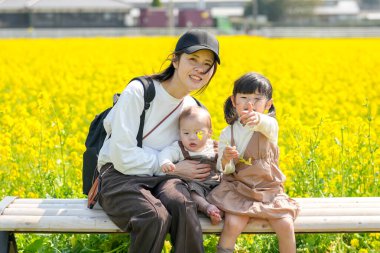 30 'lu yaşlarda Japon bir anne, kırsal bir tarlada çiçek açan bir bankta, 6 aylık bebeğini kucağında, 3 yaşındaki ikiz kuyruklu kızının yanında oturuyor. Mart ayı başlarında yakalandı..