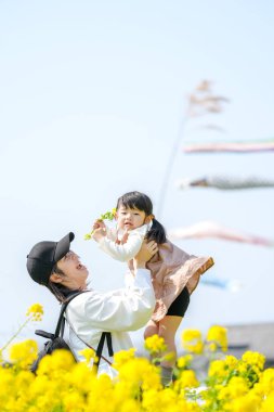 30 'lu yaşlarda Japon bir anne, kırsalda açan kolza çiçeği çiçekleriyle çevrili, sazan sazanı şeritlerinin önünde iki at kuyruklu üç yaşındaki kızını havaya kaldırır. Mart ilkbaharı.