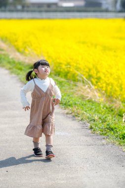 Huzurlu bir kırsal alanda, ikiz at kuyruklu 3 yaşındaki Japon bir kız Mart ayı başında çiçek açan kolza çiçeği tarlasında yürür ve baharın cazibesini somutlaştırır..