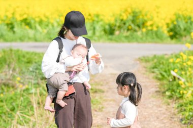 6 aylık Japon erkek çocuğu, 3 yaşındaki ikiz kuyruklu kız kardeşi ve 30 'larındaki anneleri, Mart ayında sıcak bir bahar gününde Japon kırsalında çiçek açan bir kolza tarlasında sabun köpüklerinin tadını çıkarıyorlar.