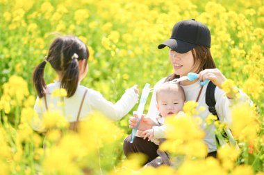 6 aylık Japon erkek çocuğu, 3 yaşındaki ikiz kuyruklu kız kardeşi ve 30 'larındaki anneleri, Mart ayında sıcak bir bahar gününde Japon kırsalında çiçek açan bir kolza tarlasında sabun köpüklerinin tadını çıkarıyorlar.