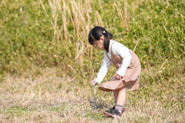 Japonya 'nın kırsal kesiminde, kır çiçekleriyle çevrili çimenli bir arazide oynayan ve Mart ayı sonlarında huzurlu bir öğleden sonra yumuşak bahar güneşinde yıkanan 3 yaşındaki Japon bir kız çocuğu..