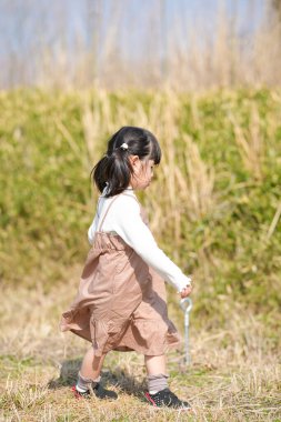 Japonya 'nın kırsal kesiminde, kır çiçekleriyle çevrili çimenli bir arazide oynayan ve Mart ayı sonlarında huzurlu bir öğleden sonra yumuşak bahar güneşinde yıkanan 3 yaşındaki Japon bir kız çocuğu..