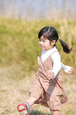 Japonya 'nın kırsal kesiminde, kır çiçekleriyle çevrili çimenli bir arazide oynayan ve Mart ayı sonlarında huzurlu bir öğleden sonra yumuşak bahar güneşinde yıkanan 3 yaşındaki Japon bir kız çocuğu..